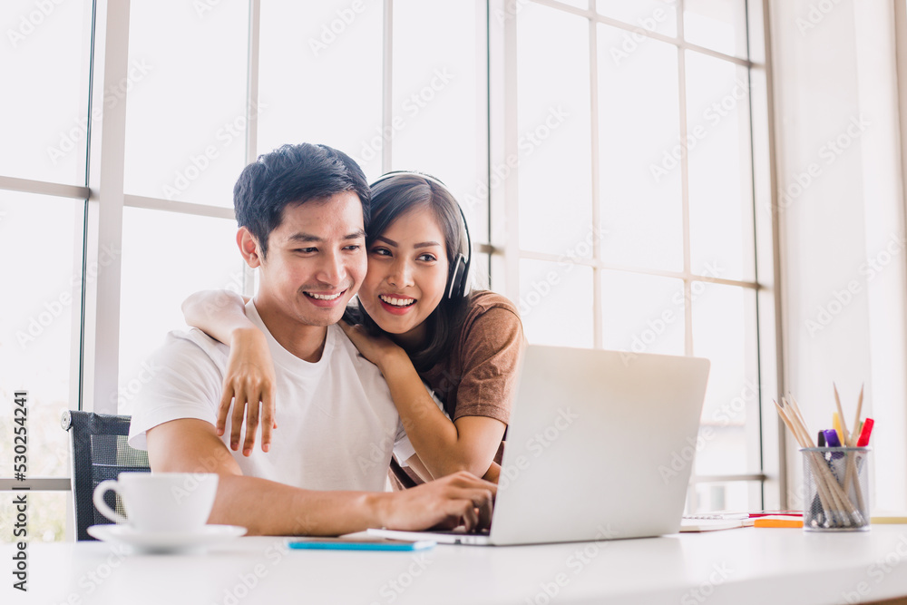 Young Asian couple playing laptop and looking at the screen at home. Cheerful couple relaxing in beautiful white room. Close up shot woman hugging her boyfriend while he working. Young Asian couple playing laptop and looking at the screen at home. Cheerful couple relaxing in beautiful white room. Close up shot woman hugging her boyfriend while he working.