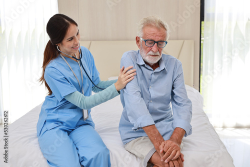 Happy caregiver consoling senior caucasian elderly man patient in bedroom at retirement house. Asian smiling nurse taking good help care and support of elder patient at house.