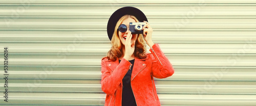 Portrait of stylish happy smiling woman with film camera taking picture wearing red jacket, black round hat on background © rohappy