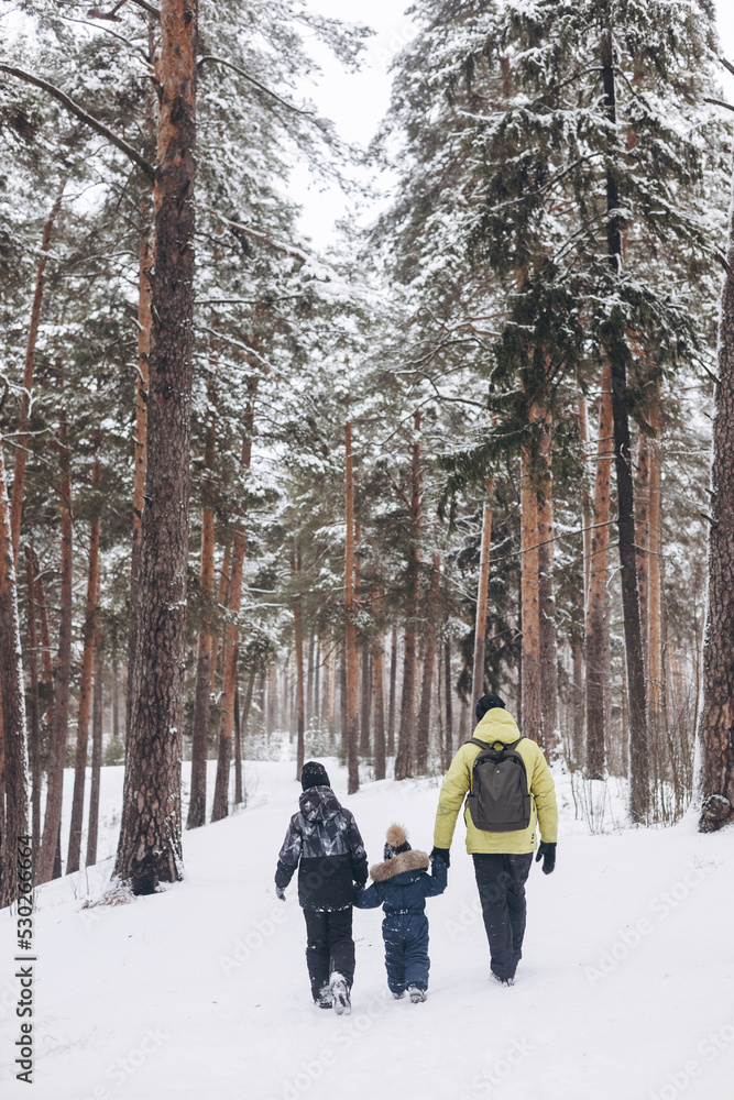 Rear view of father with backpack and little sons holding hand walking together in winter snowy forest. Wintertime activity outdoors. Concept of local travel and family weekend