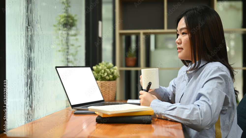 Side view asian woman employee drinking coffee at her workplace and looking through window Side view asian woman employee drinking coffee at her workplace and looking through window