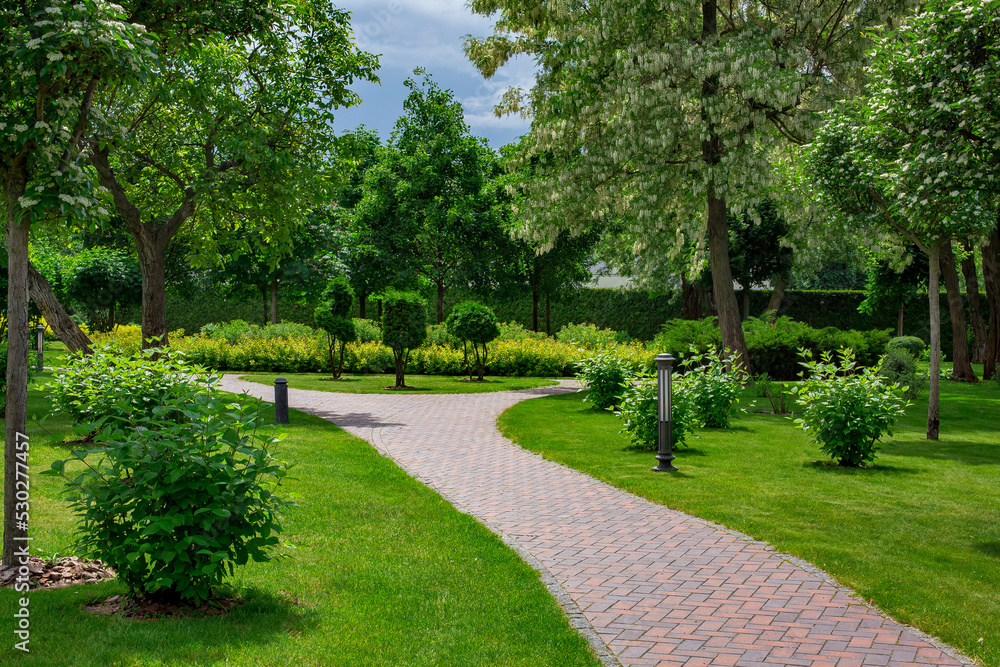 lantern iron ground garden lighting of curved park path paved with stone tiles in backyard among plants with mulching, bushes and trees surrounded by a green lawn on a sunny spring day, nobody. lantern iron ground garden lighting of curved park path paved with stone tiles in backyard among plants with mulching, bushes and trees surrounded by a green lawn on a sunny spring day, nobody.