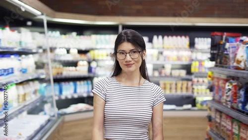 Portrait of attractive young asian woman with cart standing in supermarket with shelves of dairy on background, looking at camera and smiling. Trade business and people concept photo