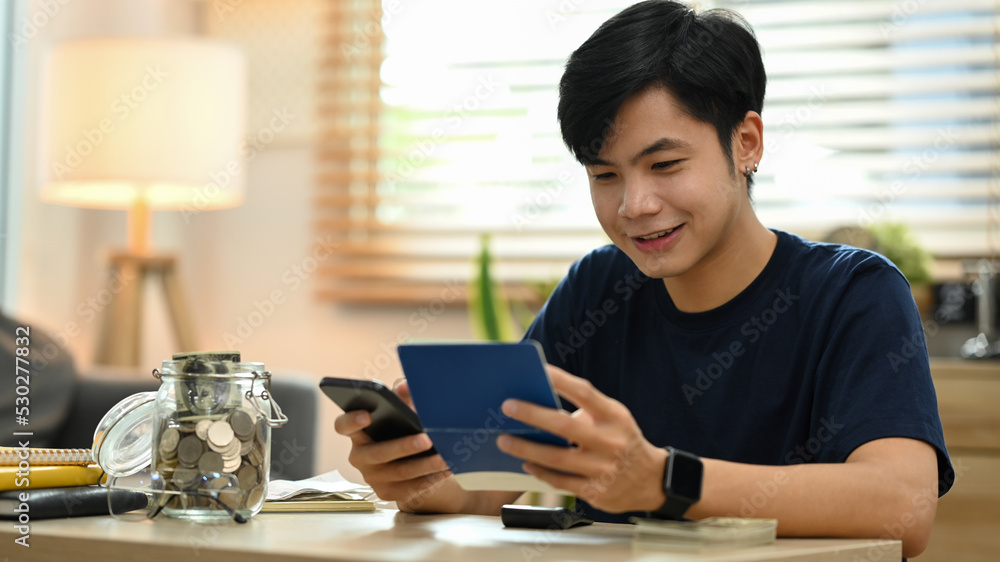 Happy young asian man using smart phone for making online payments, managing expenses finances in living room Happy young asian man using smart phone for making online payments, managing expenses finances in living room