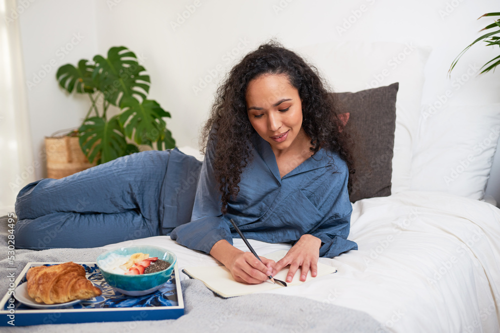 A young woman has a self-care day in bed eating breakfast and journalling