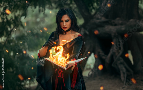 Fototapet Fantasy halloween woman witch holds old burning magic book in hand, reads spell Paper page in bright flame fire light