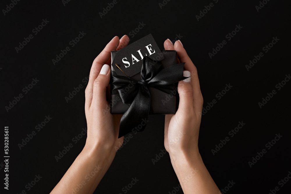 Fototapeta premium Top view photo of female hands holding black gift box with black satin ribbons and text on sale tag on isolated black background. Black Friday Sale template.