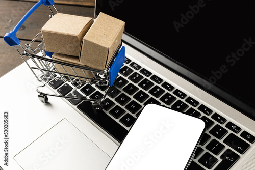 Composition of smartphone, shopping cart with boxes and laptop on wooden background