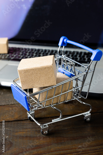 Composition of shopping cart with boxes and laptop on wooden background
