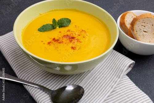 Horizontal image of bowl of carrot soup with garnish, napkin, spoon and sliced bread on slate