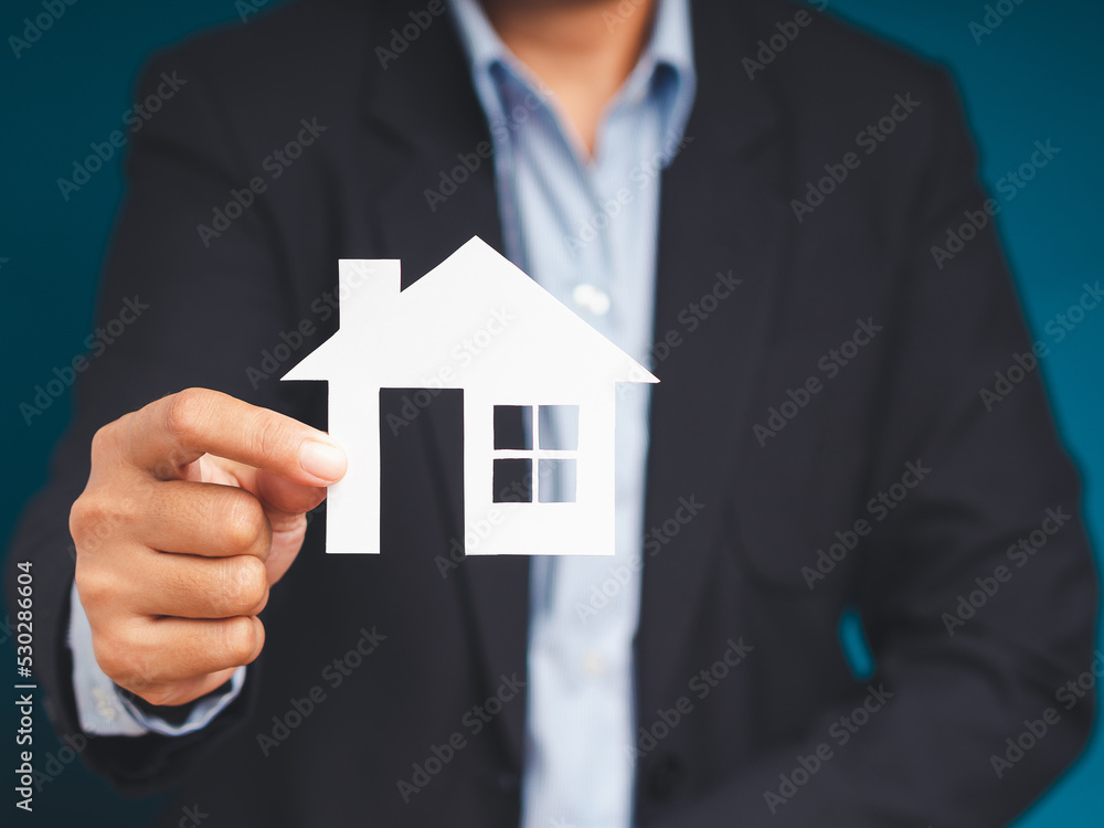Hand businessman holding a house mockup paper while standing on a blue background Hand businessman holding a house mockup paper while standing on a blue background
