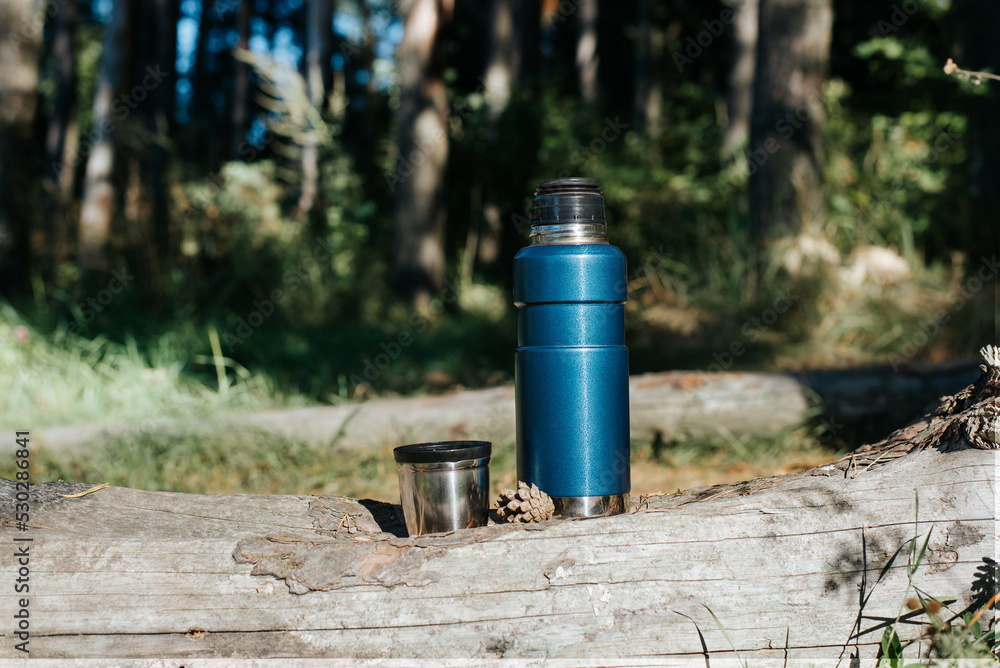 A vacuum camping thermos and a mug standing on a log in the forest on a sunny day A vacuum camping thermos and a mug standing on a log in the forest on a sunny day