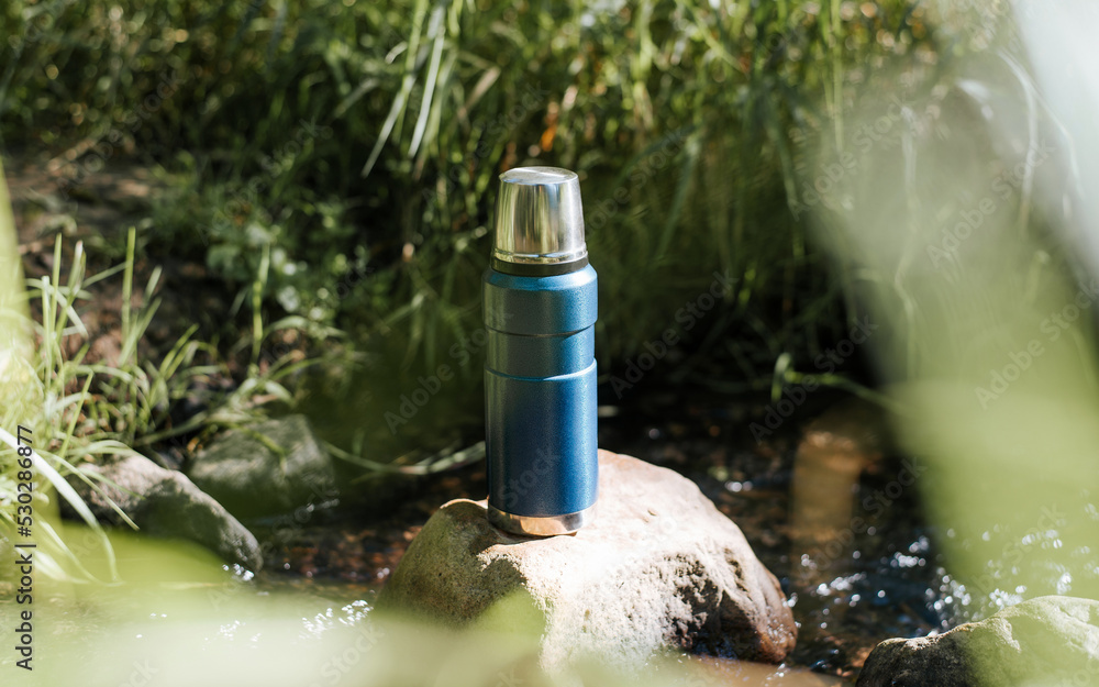 Close-up of a vacuum thermos, a camping flask on a stone near a stream in the forest on a sunny day Close-up of a vacuum thermos, a camping flask on a stone near a stream in the forest on a sunny day