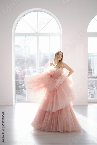 A girl in a beautiful lush long dress of dusty rose color stands near an open window. Vintage bridal glamor. Fashion shooting in a wedding or evening dress