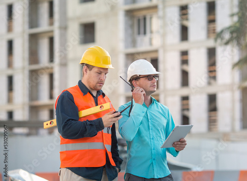 young men in a shirt and yellow and white construction helmets are standing at a construction site. They have walkie-talkies and a tablet.