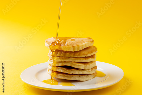 Horizontal image of maple syrup pouring onto stack of pancakes, yellow background and copy space