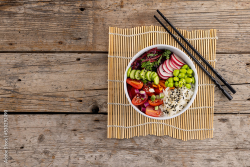 Overhead view of hawaiian poke bowl with chopsticks and copy space on mat and wooden background