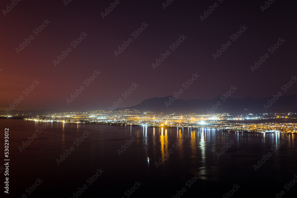Landscape of sea, sea shore with city lights, mountains, cloudless sky at night and horizon