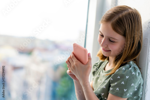 Free Download Pretty preteen girl looking at smartphone and smiling at home close to window with daylight. Beautiful kid with cell phone indoors - GFXLoad