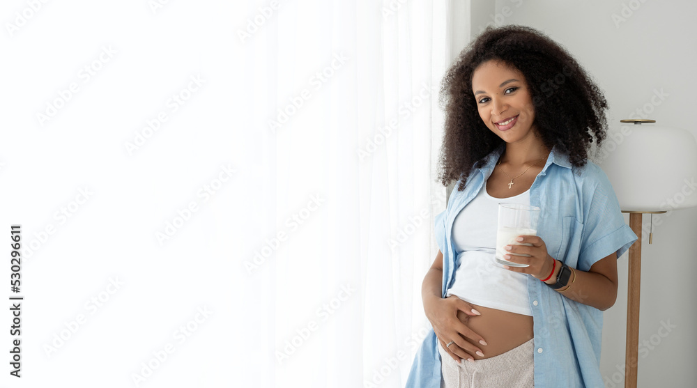 Black pregnant woman drinking milk, smile at camera, stroking tummy near window at home. Healthy eating for pregnancy, balance and vitamins. Banner Black pregnant woman drinking milk, smile at camera, stroking tummy near window at home. Healthy eating for pregnancy, balance and vitamins. Banner