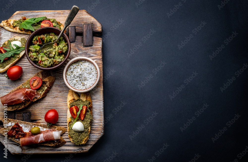 Set of bruschettas with jamon, olives, pesto, tomatoes and basil served on wooden board with guacamole and salt with herbs. Food composition with mediterranean antipasti on background with copy space Set of bruschettas with jamon, olives, pesto, tomatoes and basil served on wooden board with guacamole and salt with herbs. Food composition with mediterranean antipasti on background with copy space