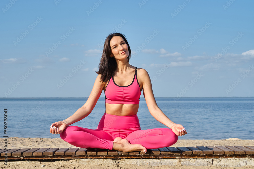 Women in sportswear sitting in a lotus position and meditating during yoga classes on the seashore. Practicing yoga lesson, breathing, meditation, doing Ardha Padmasana exercises. Wellness concept
