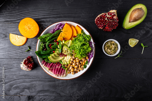 Fresh salad with roasted chickpeas, avocado, persimmon, spinach, avocado, watermelon radish and seeds on a dark background. Long banner format. top view