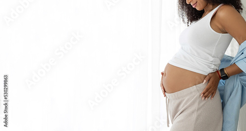 Close up of happy Afro-American pregnant woman with beautiful belly near window at home. Pregnancy, motherhood, future parent, childbirth and expectation concept - pregnancy, motherhood, expectation