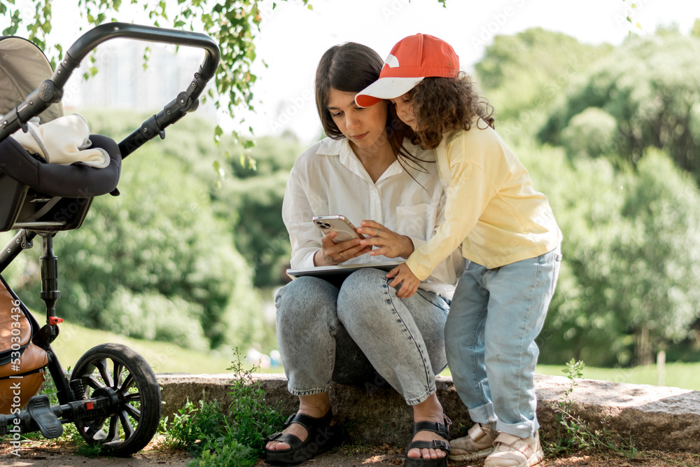 A young businesswoman with her little daughter and a baby in a stroller is using a smartphone in a park on a summer day.Remote work, freelance,working moms,online education.