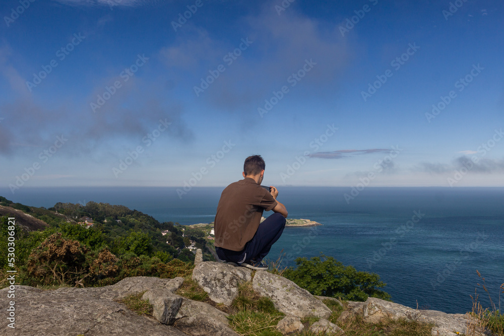 Young man sits on top of a rocky hill in Killiney and watches the sea on a gorgeous sunny day, Killiney, Ireland