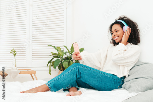 Beautiful black woman with afro curls hairstyle.Smiling model in sweater and jeans.Sexy carefree female listening music in wireless headphones in morning.Lying in bed in white interior. Taking selfie