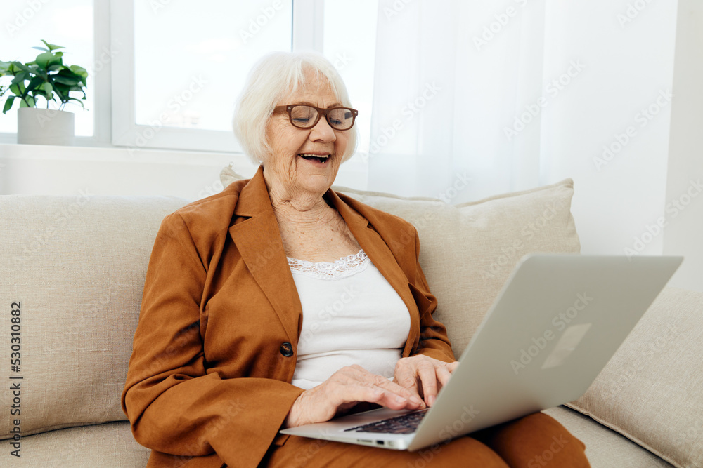 a happy elderly woman with gray white hair is sitting in a bright apartment on a beige cozy sofa happily working at a laptop