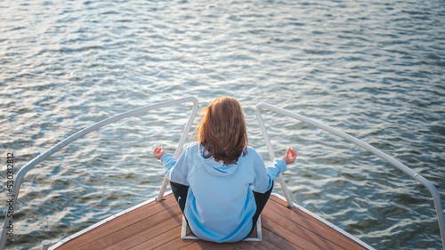 Young woman relaxing and meditating while sitting in a lotus position on the prow of a boat on the water during sunset
