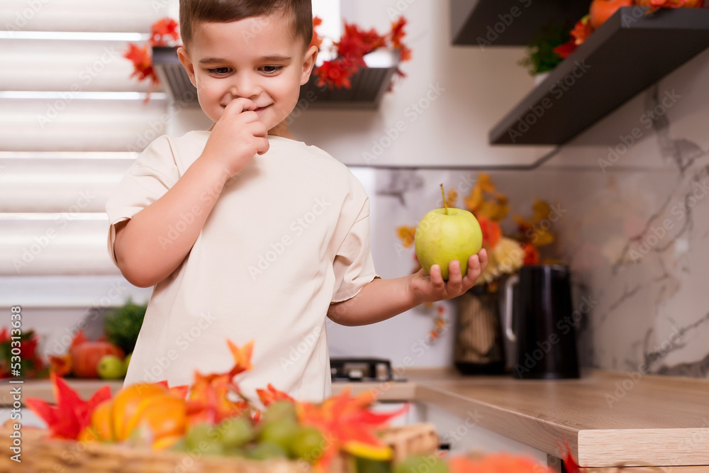 Smiling cute boy standing at counter with fruits, autumn leaves and pumpkin. Autumn decorated kitchen. Holidays concept. Smiling cute boy standing at counter with fruits, autumn leaves and pumpkin. Autumn decorated kitchen. Holidays concept.