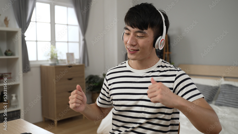 happy asian Japanese man in casual wear is sitting dancing at desk while enjoying listening to music from headphones in a bright bedroom at home. happy asian Japanese man in casual wear is sitting dancing at desk while enjoying listening to music from headphones in a bright bedroom at home.