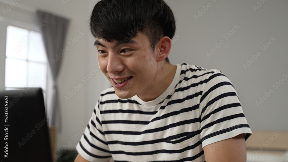 surprised asian adult Japanese student celebrating with clenched fists while receiving college acceptance email on laptop in his bedroom at home. surprised asian adult Japanese student celebrating with clenched fists while receiving college acceptance email on laptop in his bedroom at home.