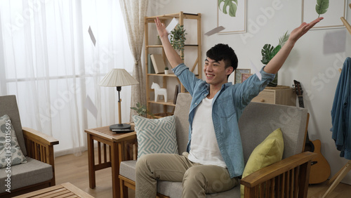 happy lucky asian Japanese man watching televised draw is throwing lottery tickets in air while celebrating with clenched fists for unexpected win in the living room at home.