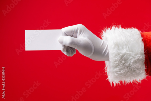 Image of hand of santa claus holding white card with copy space on red background