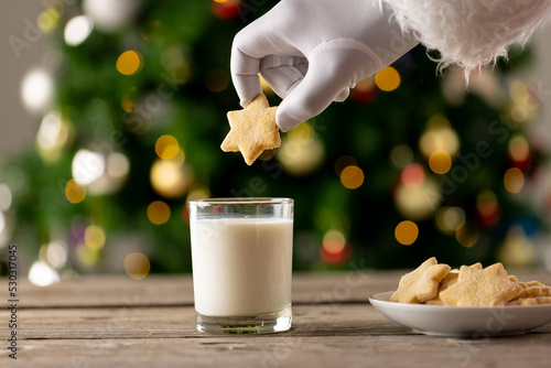Image of hand of santa claus holding christmas star biscuit over glass of milk