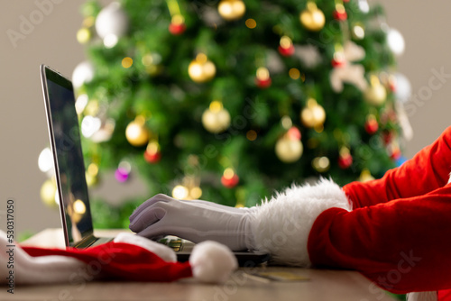 Image of hands of santa claus using laptop with christmas tree in background