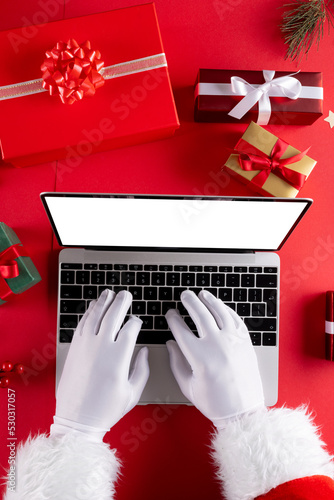 Image of hands of santa claus using laptop with blank screen and gifts on red background