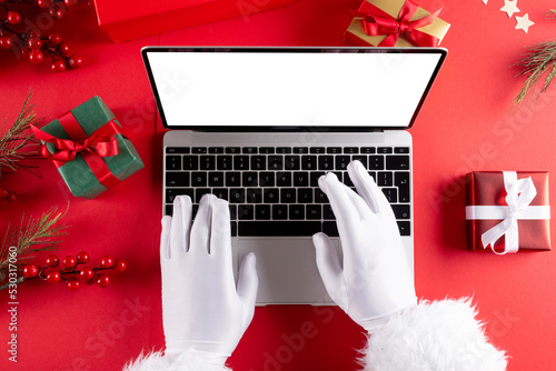 Image of hands of santa claus using laptop with blank screen and copy space on red background