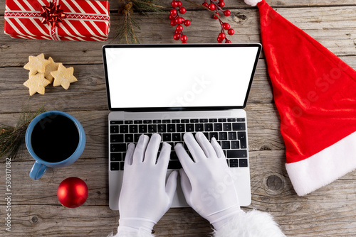 Image of hands of santa claus using laptop with blank screen and copy space on wooden background