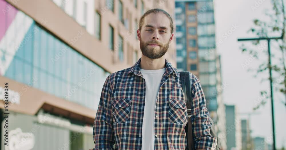 Portrait shot of young Caucasian handsome smiled man with beard and hair in tail. Outdoors. Good-looking male in motley shirt and with backpack standing outside at street. Portrait shot of young Caucasian handsome smiled man with beard and hair in tail. Outdoors. Good-looking male in motley shirt and with backpack standing outside at street.