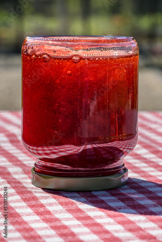 A glass jar with fresh homemade strawberry jam standing upside down on a table with a red checkered tablecloth.