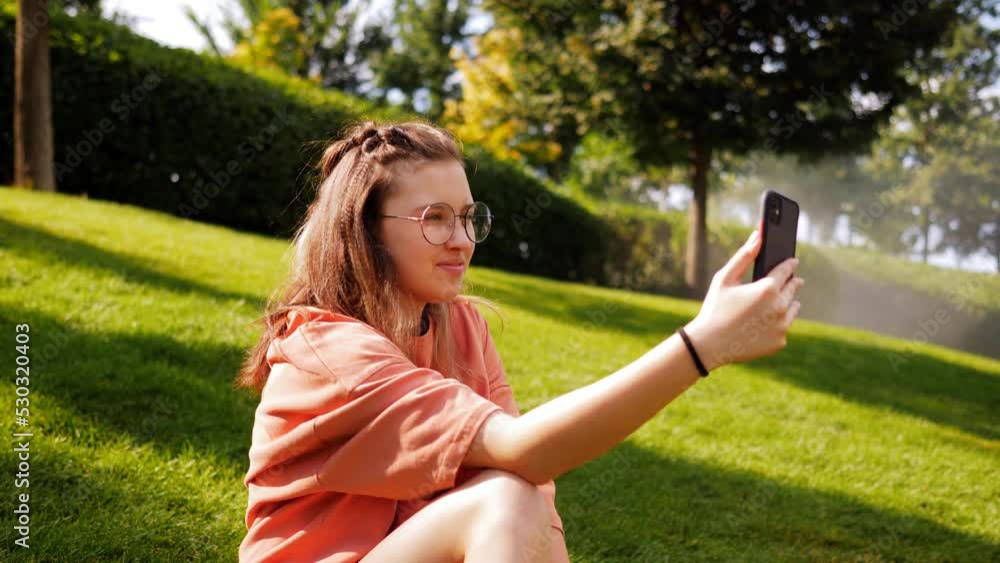 An attractive girl with glasses sitting on the grass in the park, communicating via video using a smartphone. An attractive girl with glasses sitting on the grass in the park, communicating via video using a smartphone.