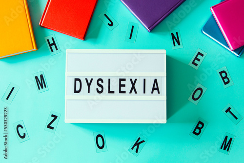 Dyslexia awareness, help children with reading, learning difficulties concept. Lightbox with DYSLEXIA word. Colorful books and different letters on light blue background. Top view