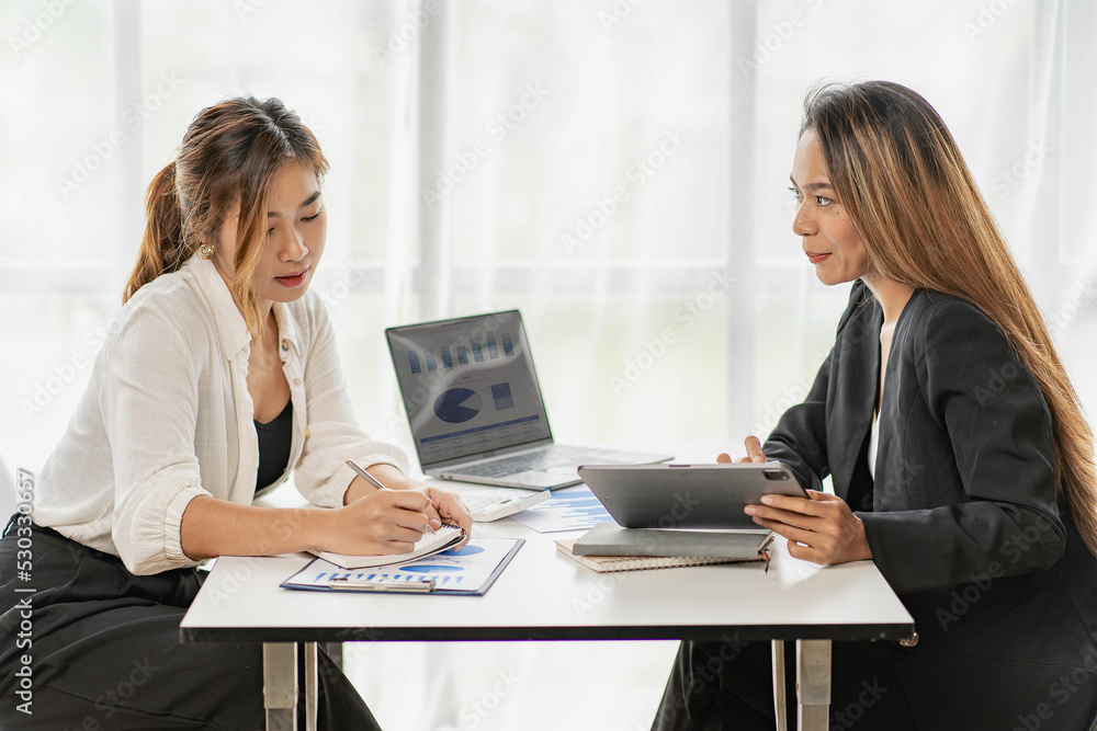 A team of Asian businesswomen discussing something and smiling as they sit at a desk in the office of women working together analyzing finances. A team of Asian businesswomen discussing something and smiling as they sit at a desk in the office of women working together analyzing finances.