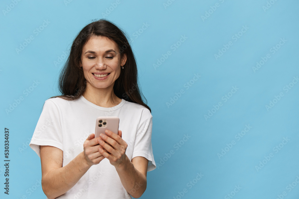 a happy, joyful woman with beautiful long hair, in a light T-shirt, stands with a fashionable phone in her hands and laughs very happily. Vertical photo on light blue background with space for text a happy, joyful woman with beautiful long hair, in a light T-shirt, stands with a fashionable phone in her hands and laughs very happily. Vertical photo on light blue background with space for text