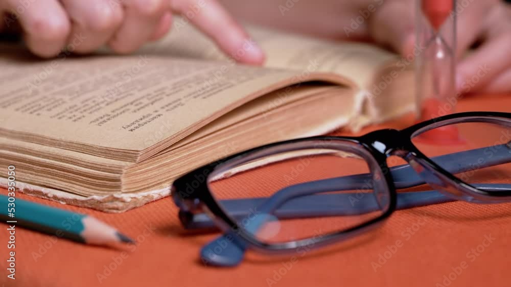 Woman Reading a Book by Candlelight in a Dark Room, Follows a Text with Finger. Close up. Girl fast runs finger over old yellow page of an old book. Hourglass. Mysterious, mystical atmosphere. Night. Woman Reading a Book by Candlelight in a Dark Room, Follows a Text with Finger. Close up. Girl fast runs finger over old yellow page of an old book. Hourglass. Mysterious, mystical atmosphere. Night.